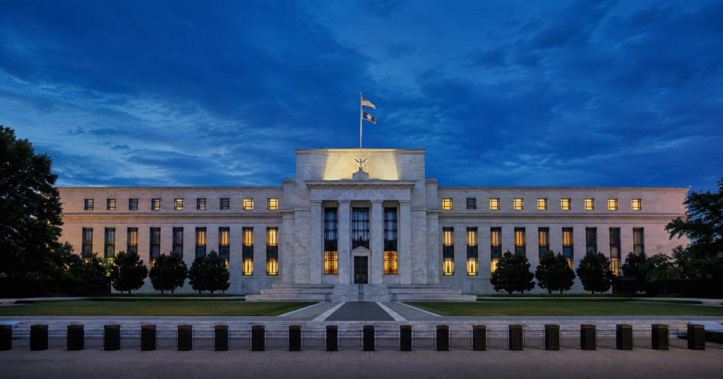 Federal Reserve Eccles Building at blue hour with warm light in a few windows beneath gathering clouds, after the July 2026 FOMC meeting's divided 9–3 vote to hold rates